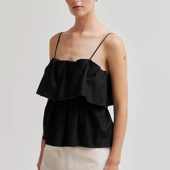 Woman wearing a black ruffled top and white pants on a plain background