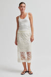 Woman wearing a white tank top and lace skirt on a plain background