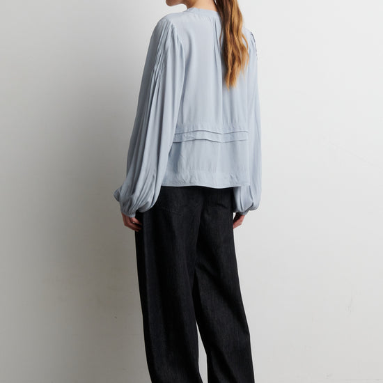 Woman wearing a light blue shirt and black pants against a white wall from Second Female