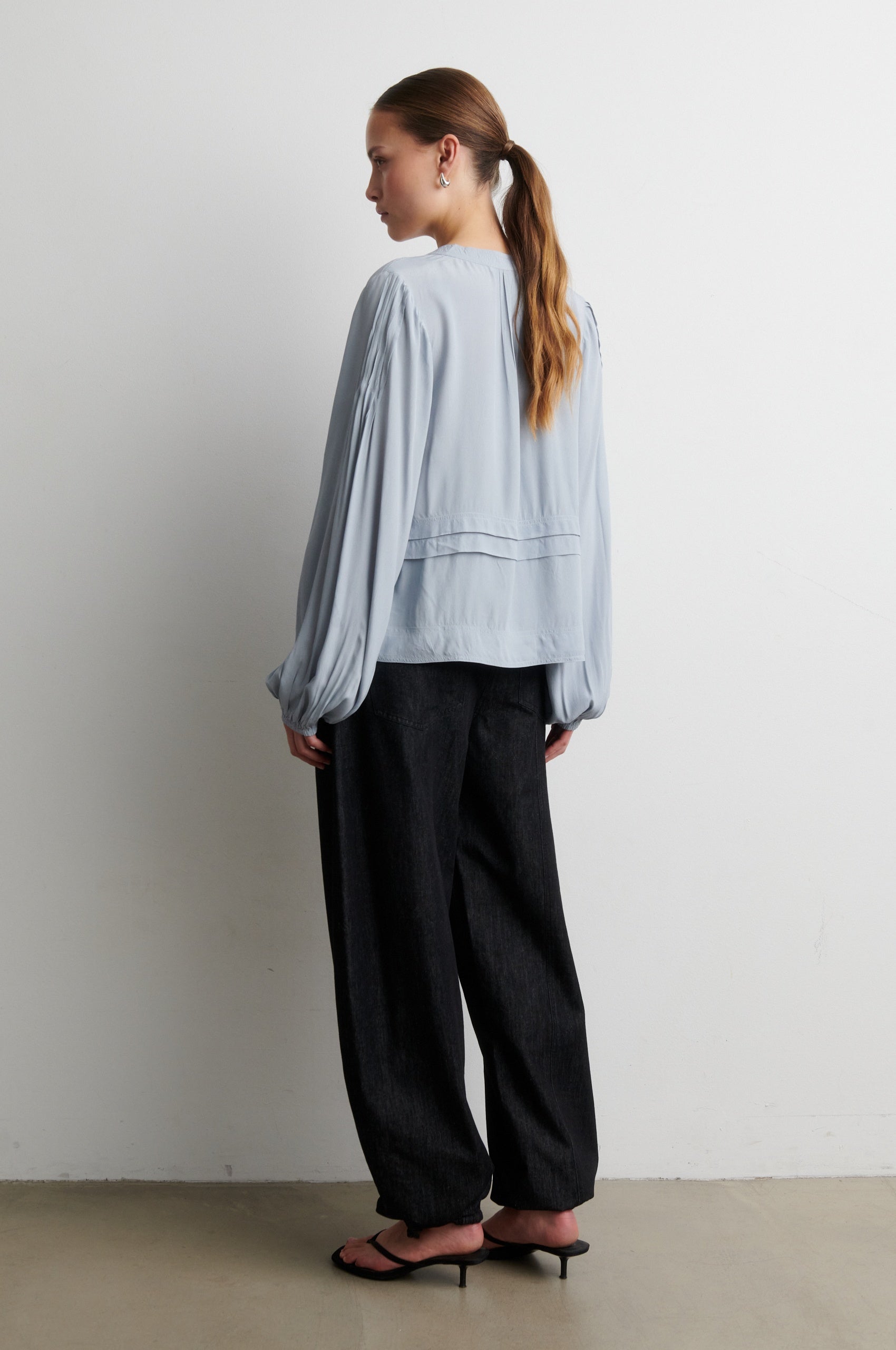 Woman wearing a light blue shirt and black pants against a white wall from Second Female