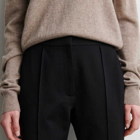 Person wearing a beige sweater and black trousers against a neutral background from Second Female