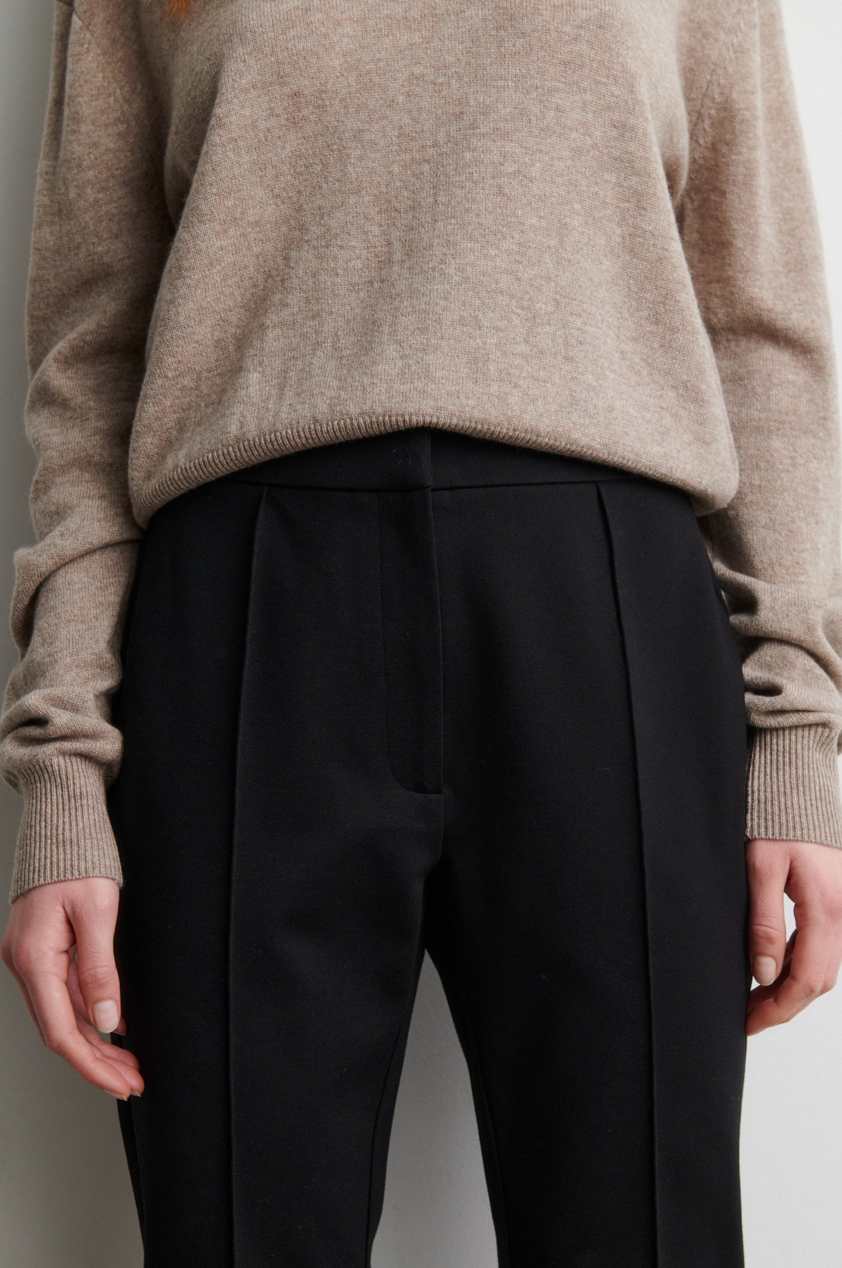Person wearing a beige sweater and black trousers against a neutral background from Second Female