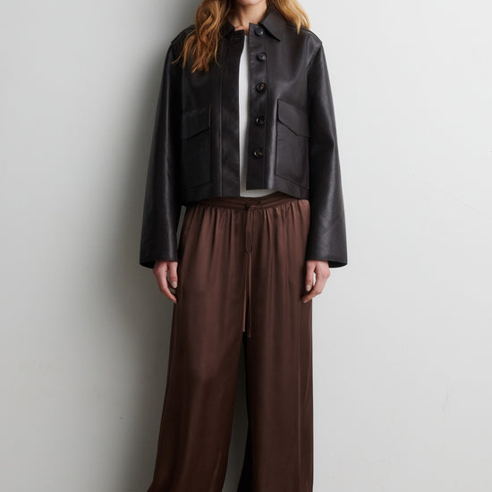 Woman wearing a black jacket and brown trousers  against a white wall from Second Female