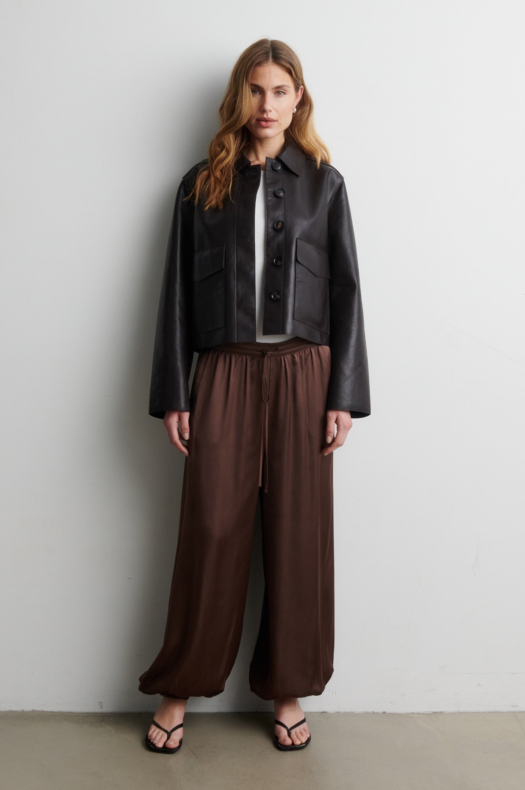 Woman wearing a black jacket and brown trousers  against a white wall from Second Female