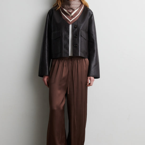 Woman wearing a black jacket and brown pants against a white wall from Second Female