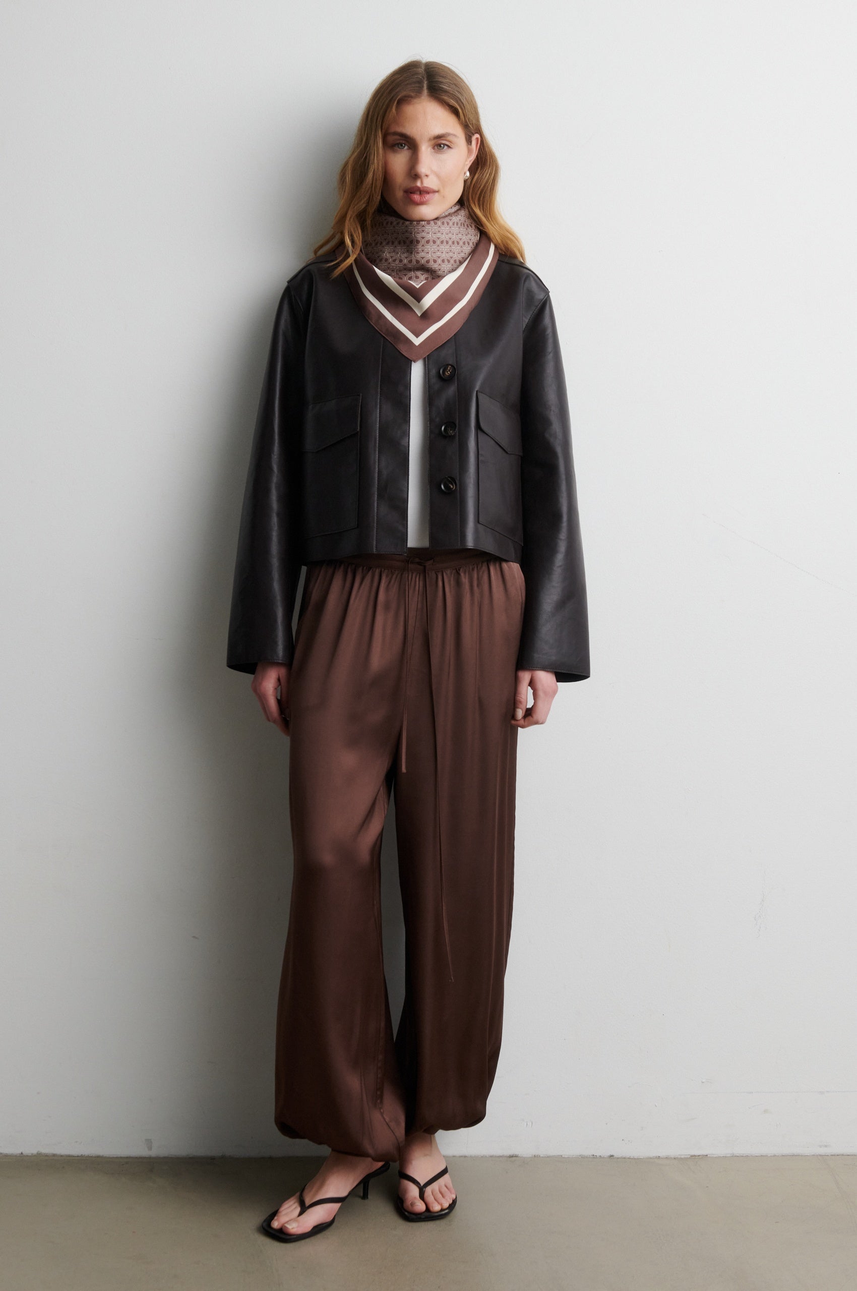 Woman wearing a black jacket and brown pants against a white wall from Second Female
