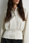 A close-up of a model wearing a cream-colored long-sleeve button-up blouse with a structured, gathered waist and black leather pants against a neutral light gray background.