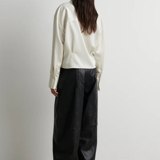 A model shown from the back wearing a cream-colored long-sleeve top and black wide-leg pants, standing against a plain light gray wall.