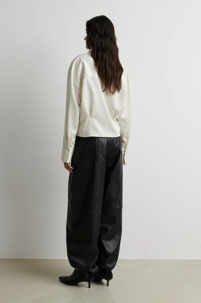 A model shown from the back wearing a cream-colored long-sleeve top and black wide-leg pants, standing against a plain light gray wall.