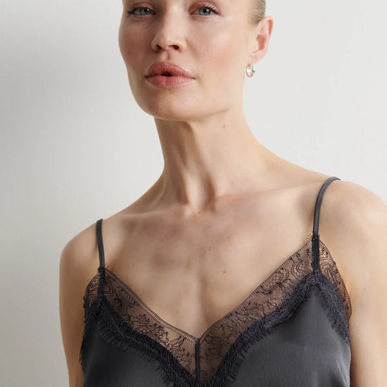 A woman wearing a dark gray satin camisole with black lace trim and thin spaghetti straps, photographed against a neutral light gray background.