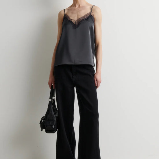 A model stands against a plain off-white wall wearing a dark gray satin camisole with black lace trim at the neckline, paired with wide-leg black trousers and black pointed-toe heels. She holds a small black handbag.