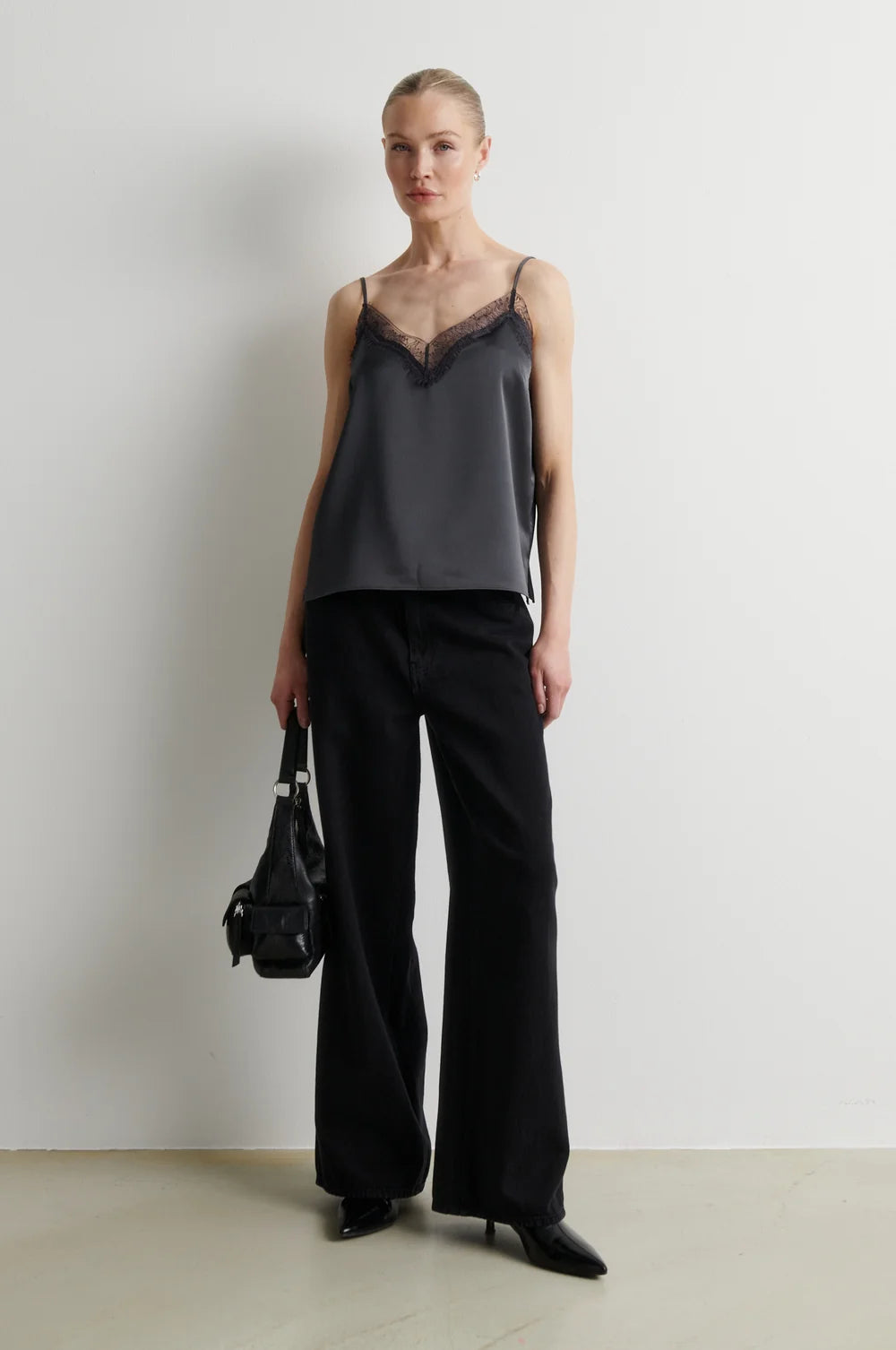 A model stands against a plain off-white wall wearing a dark gray satin camisole with black lace trim at the neckline, paired with wide-leg black trousers and black pointed-toe heels. She holds a small black handbag.