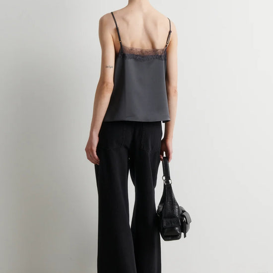 A woman stands with her back to the camera wearing a dark gray camisole with black lace trim and black wide-leg trousers, holding a black handbag against a plain light gray studio background.