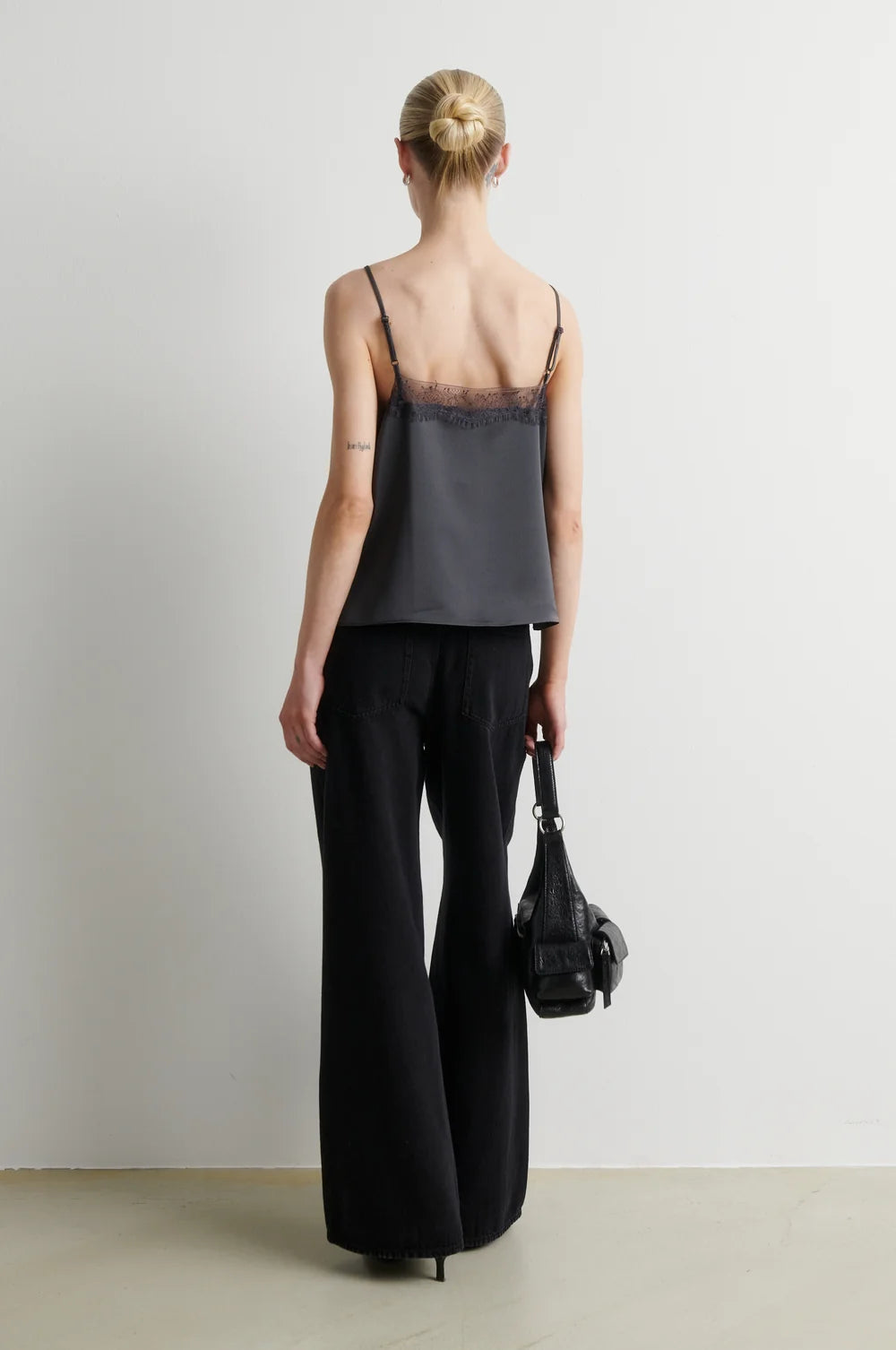 A woman stands with her back to the camera wearing a dark gray camisole with black lace trim and black wide-leg trousers, holding a black handbag against a plain light gray studio background.