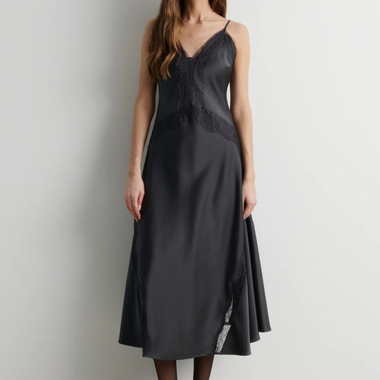 A woman modeling a dark charcoal gray midi slip dress with thin straps, a lace-trimmed V-neckline, and a flared skirt, paired with black tights and black heels against a plain light gray wall.