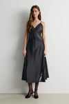A woman modeling a dark charcoal gray midi slip dress with thin straps, a lace-trimmed V-neckline, and a flared skirt, paired with black tights and black heels against a plain light gray wall.