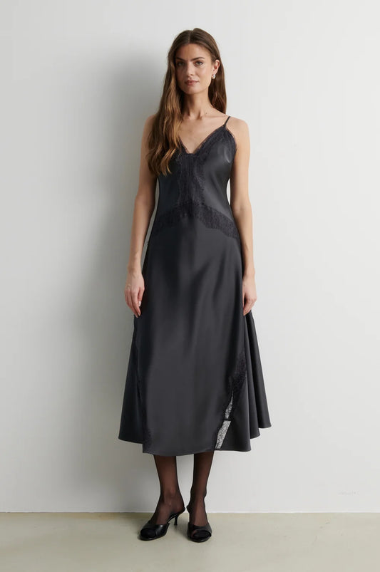 A woman modeling a dark charcoal gray midi slip dress with thin straps, a lace-trimmed V-neckline, and a flared skirt, paired with black tights and black heels against a plain light gray wall.