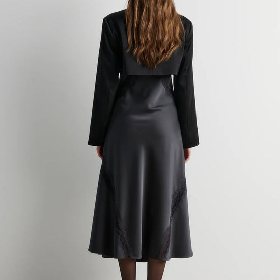 A woman viewed from behind wearing a black long-sleeve cropped jacket and a black satin midi skirt with subtle tonal embroidery, standing against a plain light gray wall.