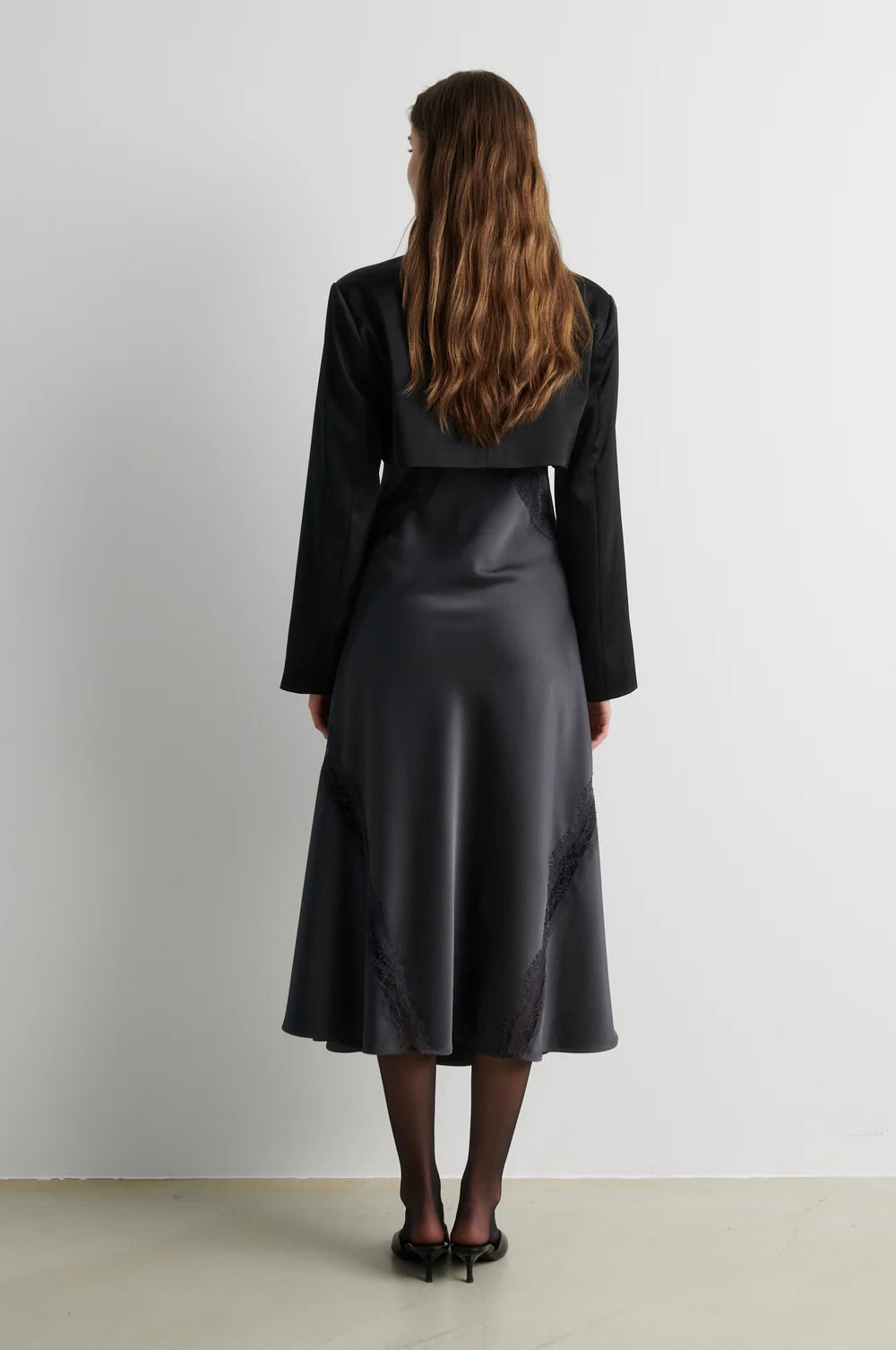 A woman viewed from behind wearing a black long-sleeve cropped jacket and a black satin midi skirt with subtle tonal embroidery, standing against a plain light gray wall.
