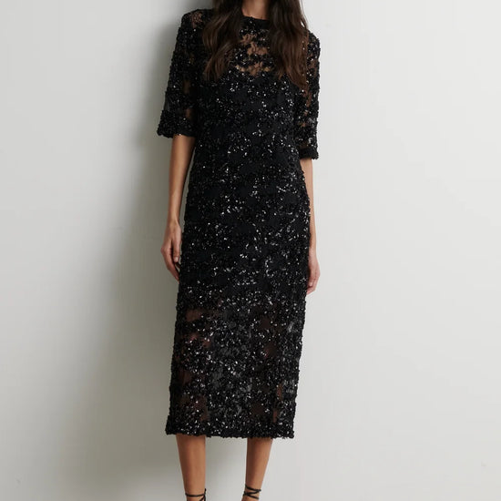 A woman modeling a black sequined midi dress with sheer three-quarter sleeves and a high round neckline, paired with black strappy high-heeled sandals, standing against a plain off-white wall.