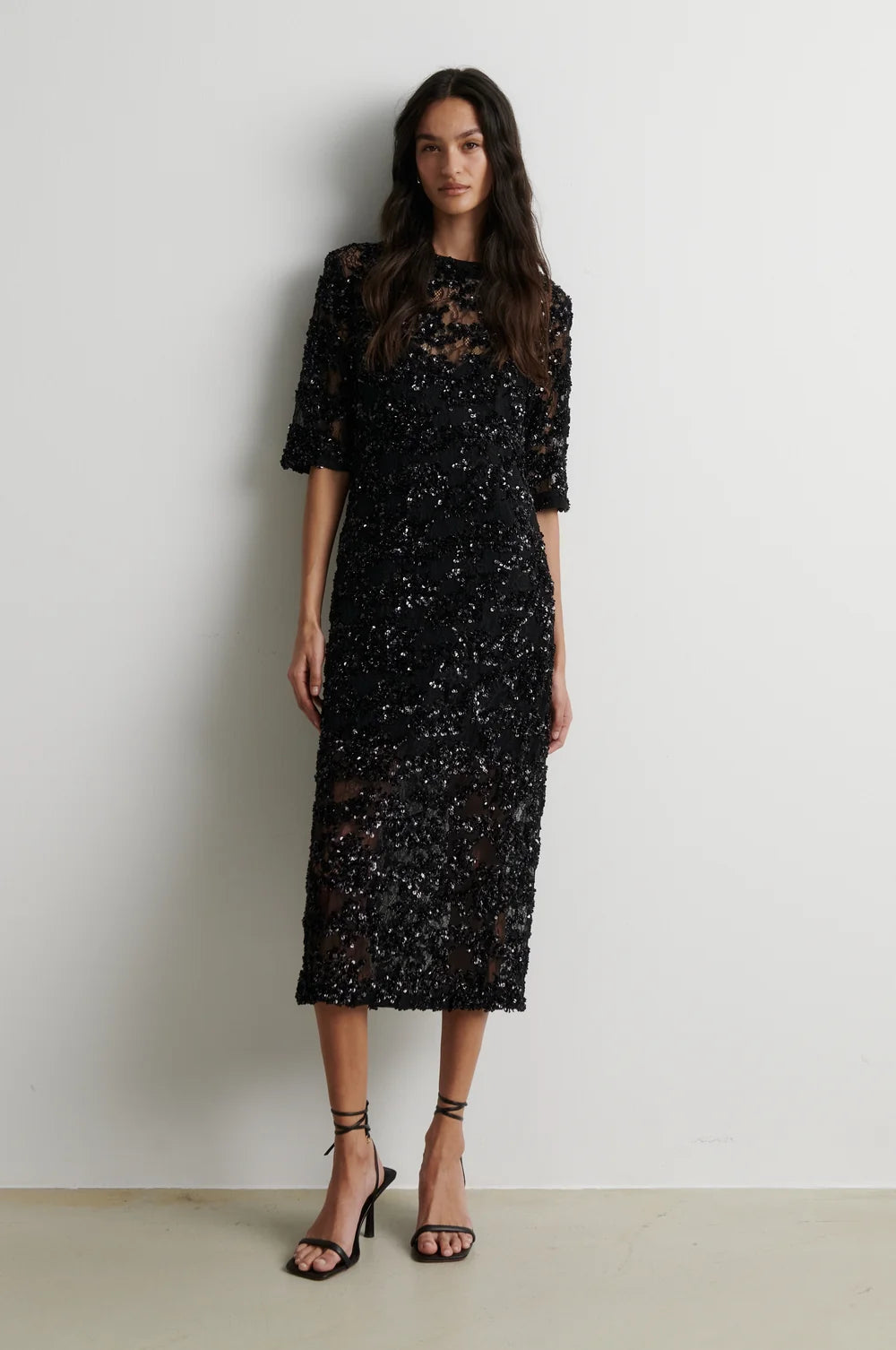 A woman modeling a black sequined midi dress with sheer three-quarter sleeves and a high round neckline, paired with black strappy high-heeled sandals, standing against a plain off-white wall.