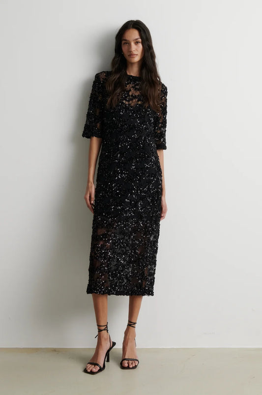 A woman modeling a black sequined midi dress with sheer three-quarter sleeves and a high round neckline, paired with black strappy high-heeled sandals, standing against a plain off-white wall.