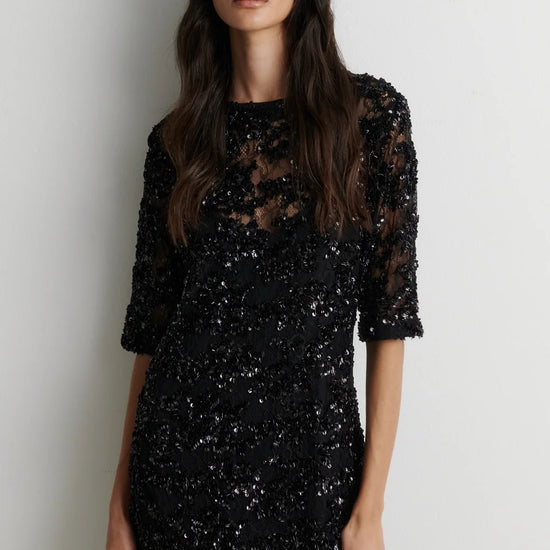 A woman modeling a black sequined midi dress with sheer three-quarter sleeves and a lace overlay, standing against a plain light gray wall.