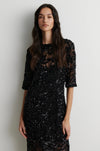 A woman modeling a black sequined midi dress with sheer three-quarter sleeves and a lace overlay, standing against a plain light gray wall.