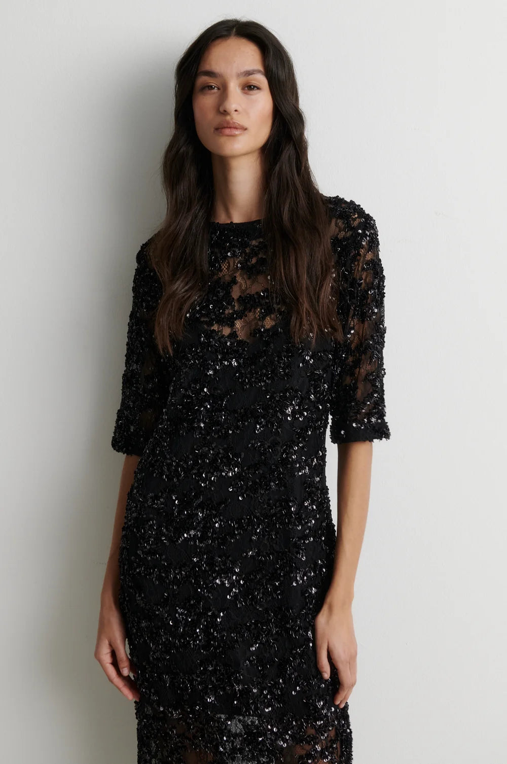 A woman modeling a black sequined midi dress with sheer three-quarter sleeves and a lace overlay, standing against a plain light gray wall.