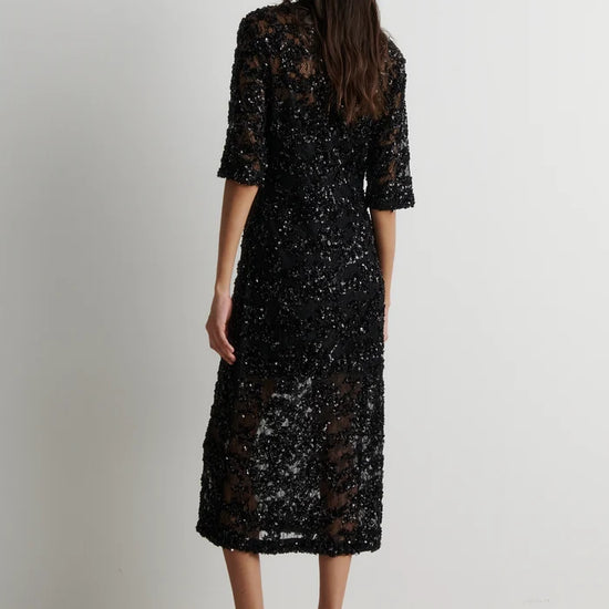 A woman shown from the back wearing a black lace midi dress with elbow-length sleeves and black strappy high-heeled sandals, standing against a plain light gray wall.
