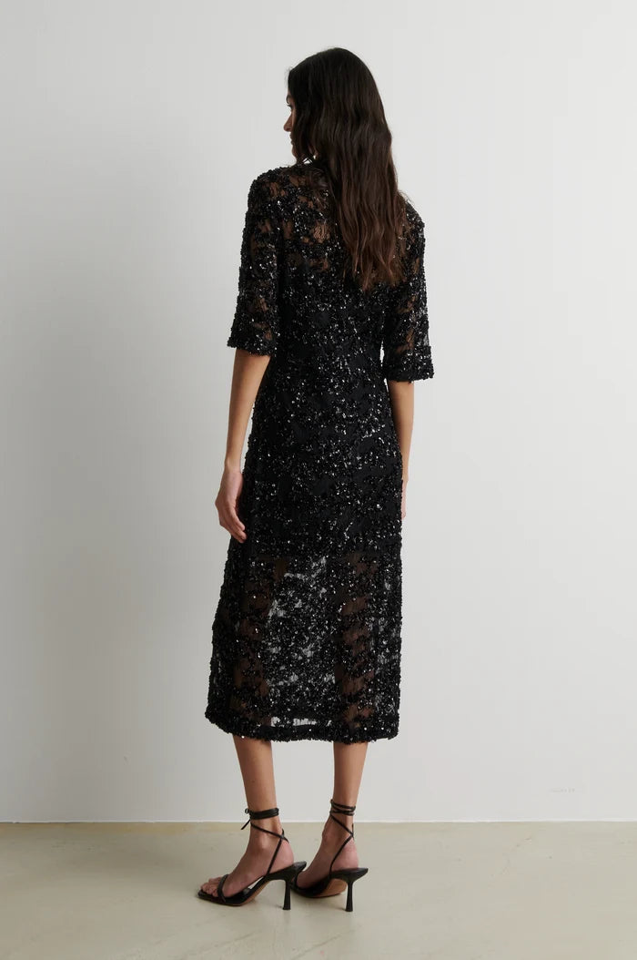A woman shown from the back wearing a black lace midi dress with elbow-length sleeves and black strappy high-heeled sandals, standing against a plain light gray wall.