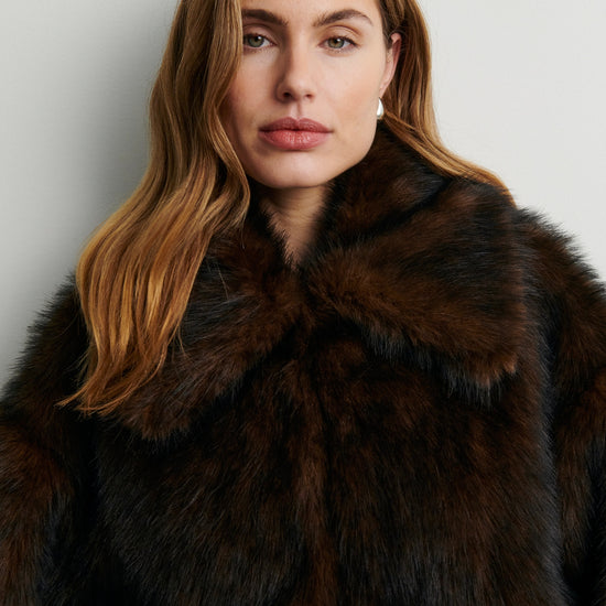 Woman wearing a dark brown fur coat against a plain background rom Second Female