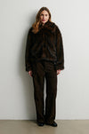 Woman wearing a dark brown fur coat and pants against a white wall from Second Female