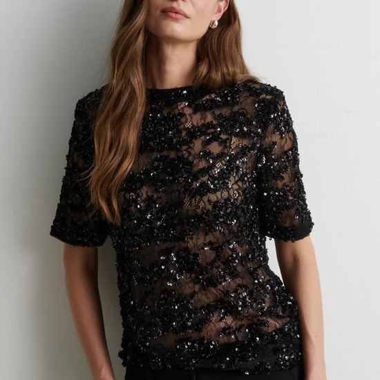 A woman wearing a sheer black short-sleeve top covered in dense sequins and lace, paired with black trousers, standing against a plain light gray wall.