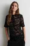 A woman wearing a sheer black short-sleeve top covered in dense sequins and lace, paired with black trousers, standing against a plain light gray wall.
