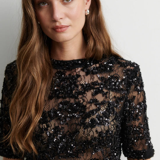 A woman wearing a black sheer lace dress with dense sequined floral detailing, photographed against a plain light gray background.