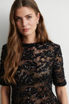 A woman wearing a black sheer lace dress with dense sequined floral detailing, photographed against a plain light gray background.