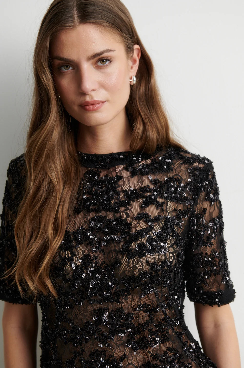 A woman wearing a black sheer lace dress with dense sequined floral detailing, photographed against a plain light gray background.