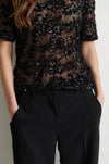 A close-up of a model wearing a black sheer lace short-sleeve top heavily embellished with sequins, paired with black trousers.