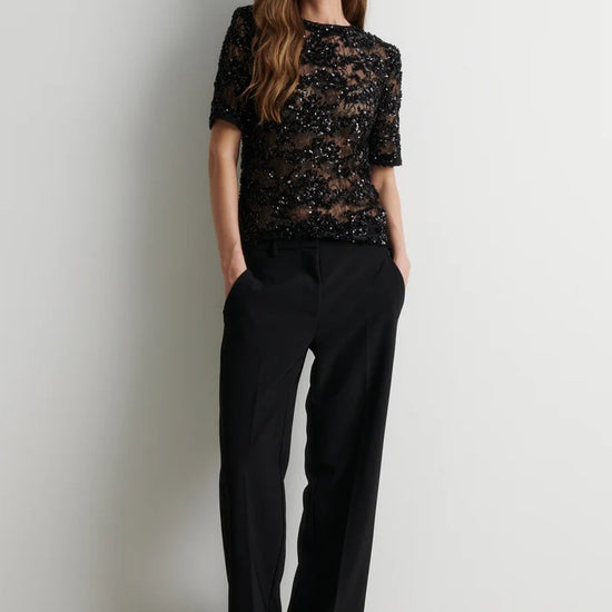 A woman stands against a plain light gray wall wearing a black short-sleeve lace top with sequined or beaded detailing and black wide-leg trousers.