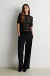 A woman stands against a plain light gray wall wearing a black short-sleeve lace top with sequined or beaded detailing and black wide-leg trousers.