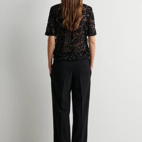 A woman viewed from behind wearing a black short-sleeve lace top and black wide-leg trousers, standing against a plain light gray wall.