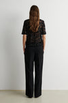 A woman viewed from behind wearing a black short-sleeve lace top and black wide-leg trousers, standing against a plain light gray wall.