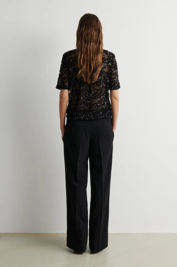 A woman viewed from behind wearing a black short-sleeve lace top and black wide-leg trousers, standing against a plain light gray wall.