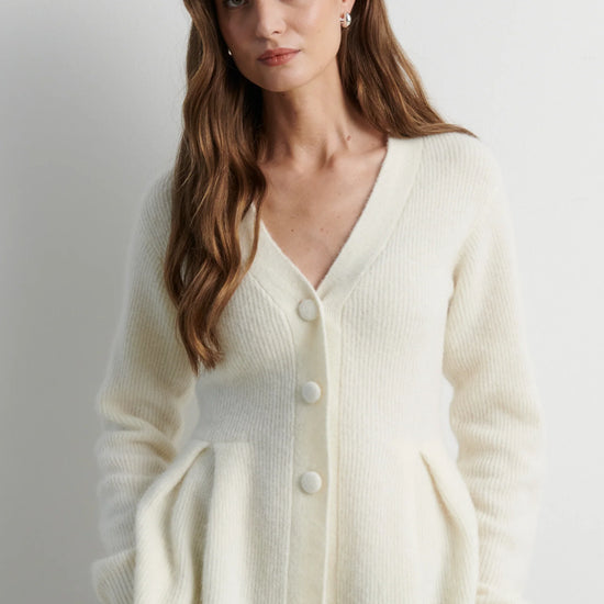 A woman wearing a cream-colored ribbed knit cardigan sweater with a V-neck and front buttons, paired with black pants, photographed against a neutral light gray background.