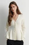 A woman wearing a cream-colored ribbed knit cardigan sweater with a V-neck and front buttons, paired with black pants, photographed against a neutral light gray background.