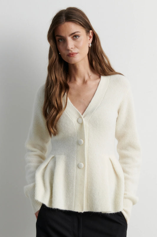 A woman wearing a cream-colored ribbed knit cardigan sweater with a V-neck and front buttons, paired with black pants, photographed against a neutral light gray background.