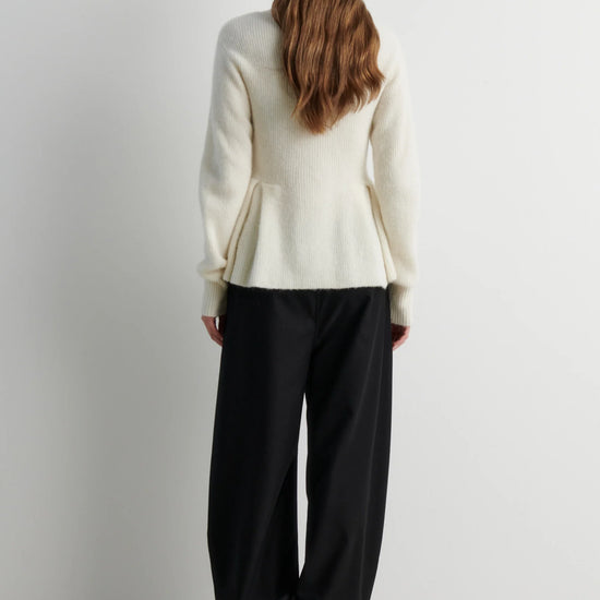 A woman viewed from behind wearing a cream-colored knit sweater and black wide-leg trousers, standing against a plain light gray wall.