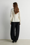 A woman viewed from behind wearing a cream-colored knit sweater and black wide-leg trousers, standing against a plain light gray wall.