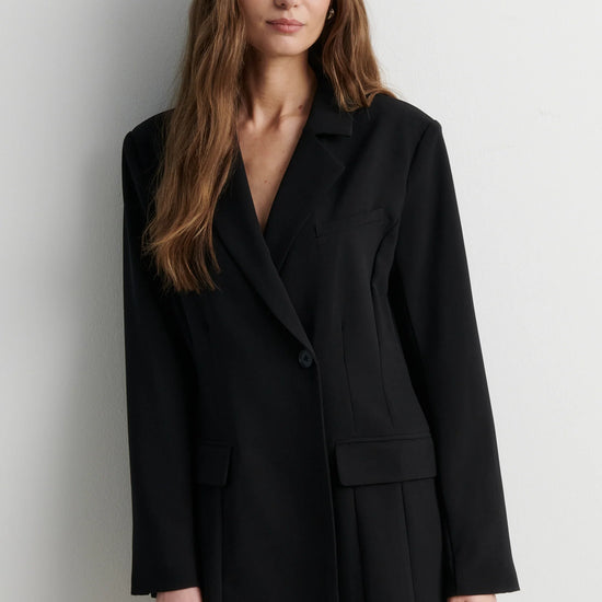 A woman modeling a black double-breasted blazer-style mini dress with a wrap front and flap pockets, photographed against a plain light gray studio background.